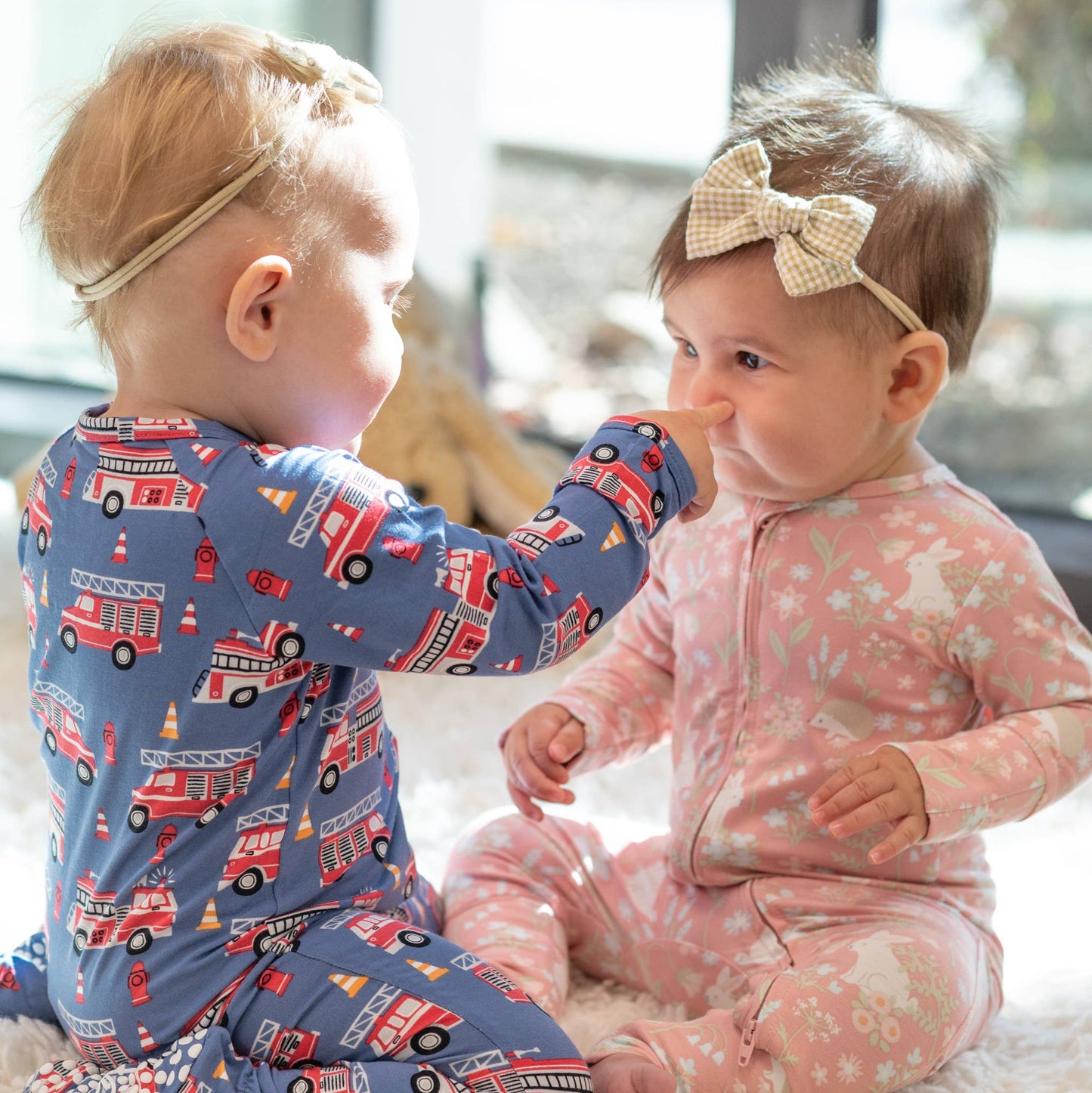 Two babies in pajamas sitting on a carpeted floor.