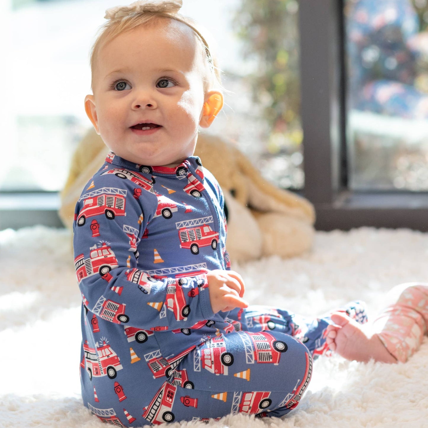Two babies sitting on a carpeted floor, one wearing pajamas with fire trucks and the other with pink pajamas.