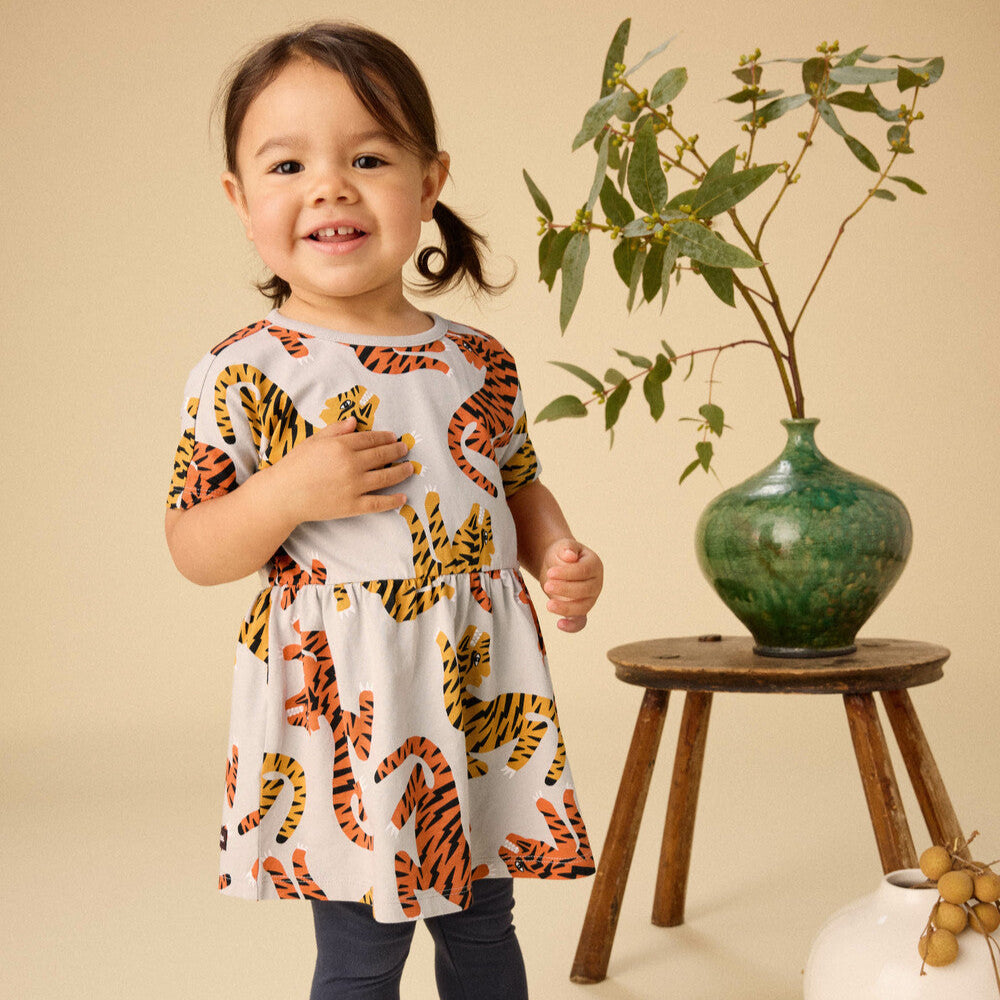 Child wearing a dress with tiger print standing next to a plant and vase on a wooden stool.